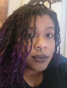 a Black woman with long locs styled up and highlighted in purple, wearing dark lipstick, a septum ring, and a lip ring, photographed in a close selfie angle. Behind her are two pieces of art depicting Black women in profile.